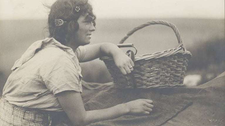 Photographic print of a Romany girl. Girl is in her teens and can seen looking towards the right of the photograph. She is leaning upon a herringbone blanket with one arm resting on a wicker basket.